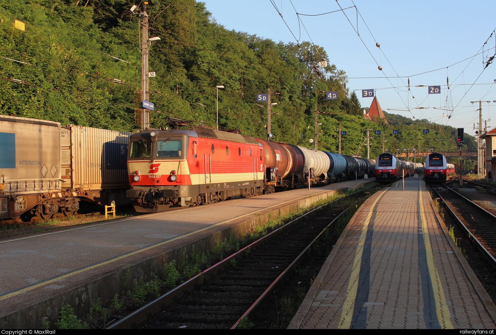 ÖBB 1144 117 in  - Die ersten Sonnenstrahlen leuchten am Morgen des 16.7.2024 über den Bahnhof Spielfeld-Straß als die 1144 117 mit einem Kesselwagenganzzug von Wien kommend in den Bahnhof rollt.
Im Bahnhof Spielfeld-Straß ist die Systemtrenndtelle zwischen den österreichischen 15kV/16,7Hz zu den slowenischen 3kV Gleichstrom und deshalb fahren die meisten Züge hier mit Schwung ein.  Südbahn | Wien Hbf -  Spielfeld Straß Railwayfans